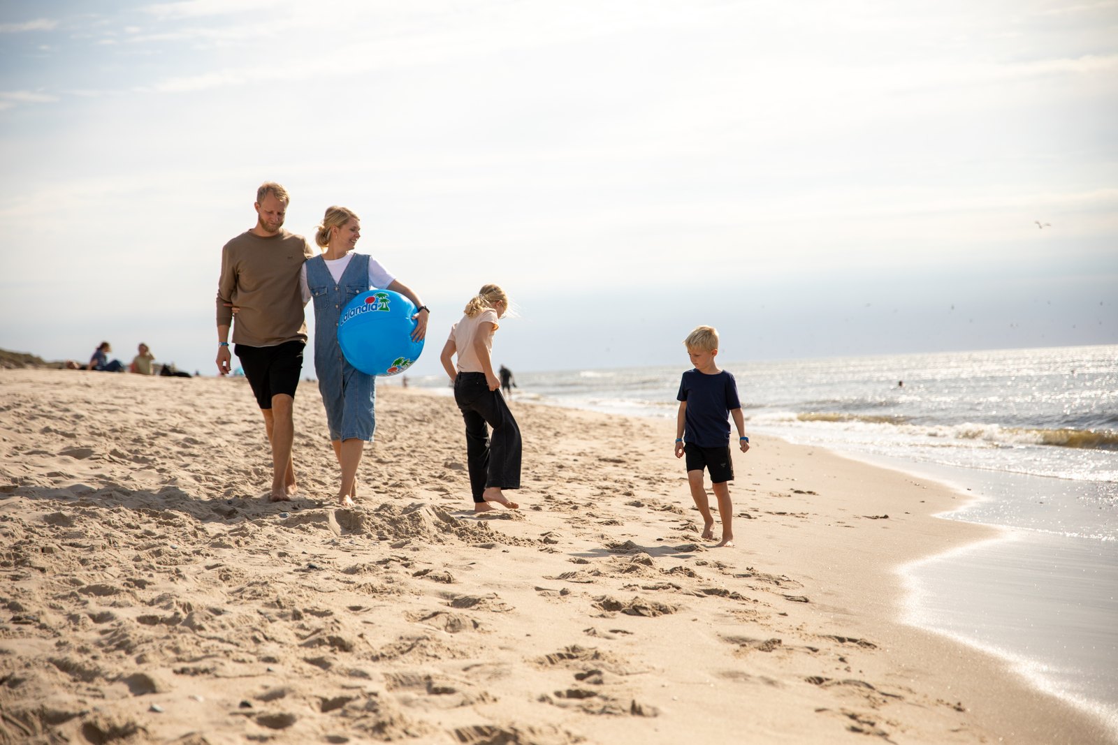 En familie med tre barn og en blå badeball går langs vannkanten på den brede sandstranda ved Søndervig, med Vesterhavet på høyre side.