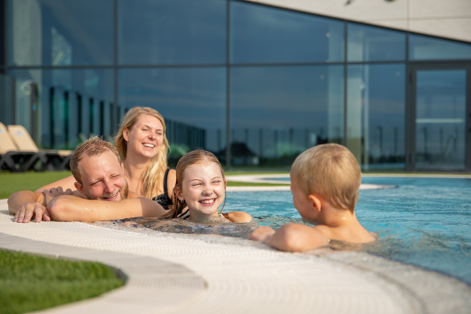 En familie med to barn ligger i kanten av det utendørs varmtvannsbassenget på Lalandia Søndervig, med glassfasaden til badelandet bak seg.