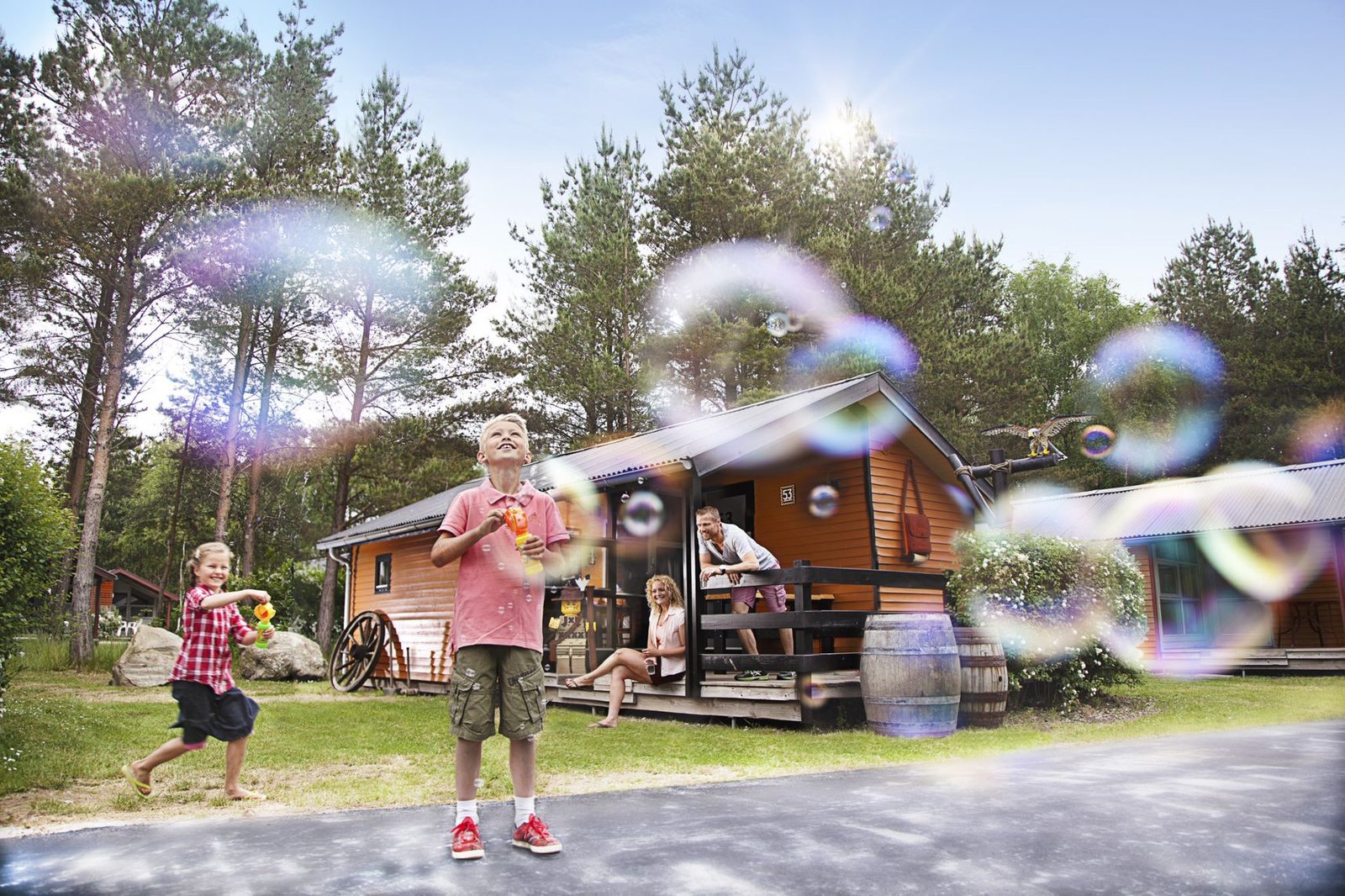 Barn blåser såpebobler foran tømmerhytter i Wild West-stil på LEGOLAND Holiday Village, med vognhjul og tønne ved terrassen og furuskog bak.