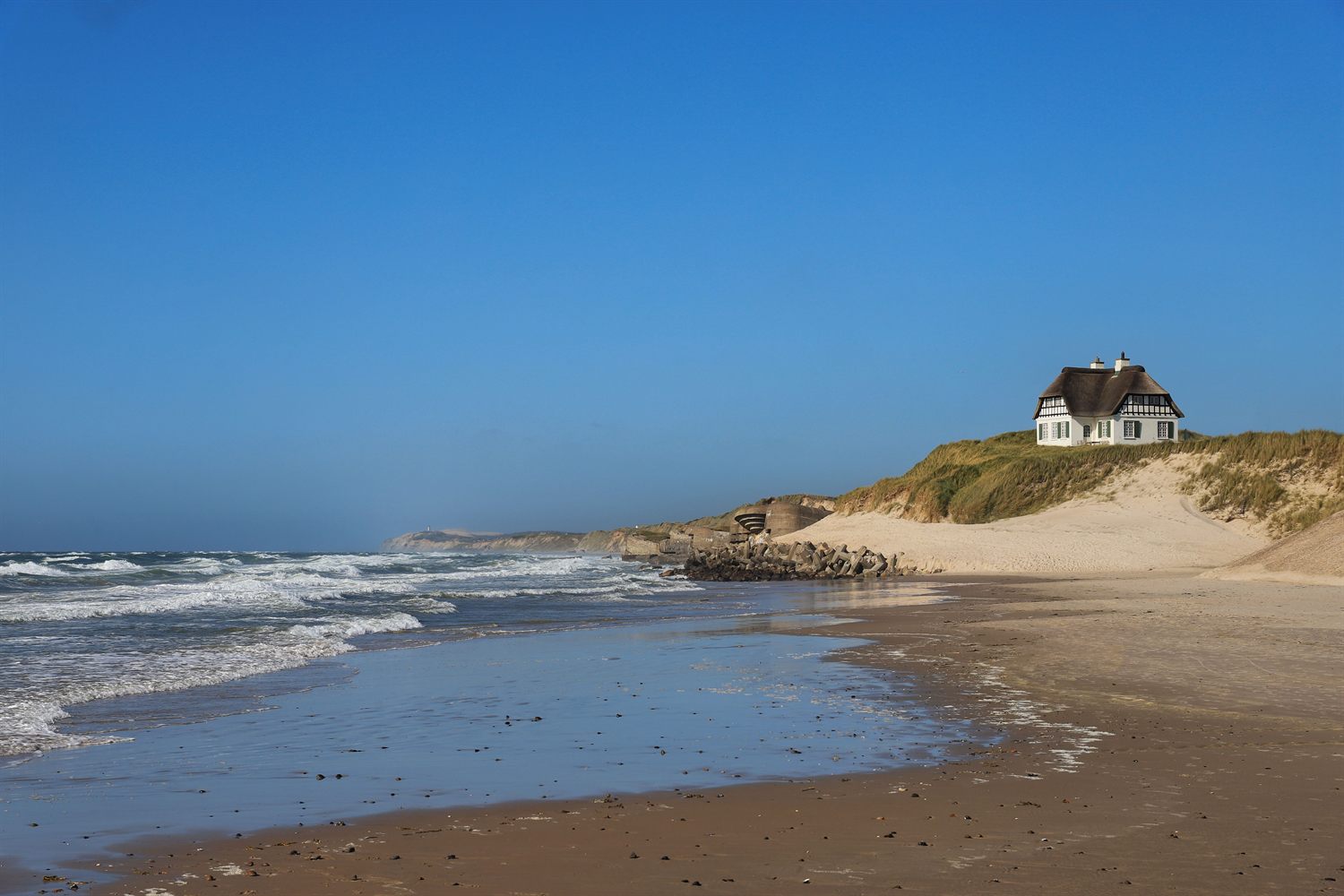 Stråtekt hvitt bindingsverkshus på en klit over en bred strand ved Vesterhavet.